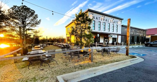 A rustic brewery with outdoor seating, surrounded by trees, at sunset. Warm light illuminates the building and patio.