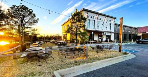 A rustic brewery with outdoor seating, surrounded by trees, at sunset. Warm light illuminates the building and patio.