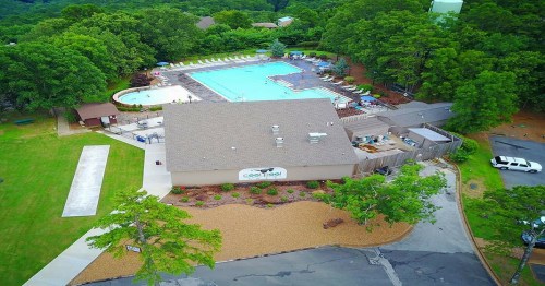 Aerial view of a pool area with lounge chairs, surrounded by trees and a parking lot. The building features a sign.