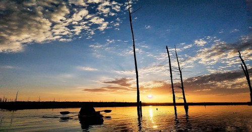 A serene sunset over a calm lake, with silhouetted trees and a person in a kayak enjoying the view.