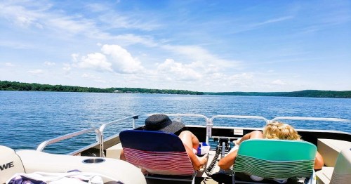 Two people relax on a boat, enjoying a sunny day on the water with blue skies and distant hills.