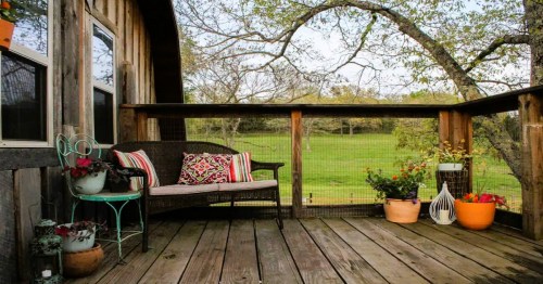 Cozy outdoor deck with a bench, colorful cushions, and potted plants, surrounded by greenery and trees.