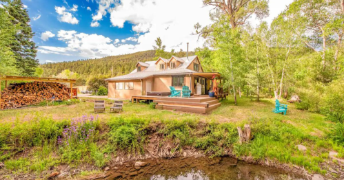 A cozy cabin by a stream, surrounded by greenery and mountains, with a deck and outdoor seating.