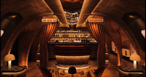 A stylish bar interior with warm lighting, curved wooden architecture, and a well-stocked shelf of bottles in the background.