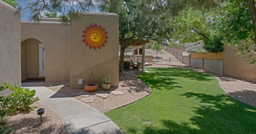 A sunny backyard with a pathway, green lawn, and a decorative sun on the wall of a beige house.