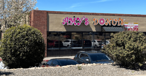A storefront of "Amy's Donuts" with a sign indicating it's open, surrounded by shrubs and parked cars.
