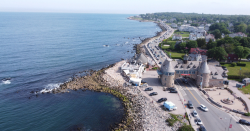 Aerial view of a coastal road with rocky shoreline, buildings, and a calm blue ocean under a clear sky.