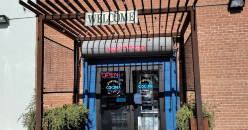 Entrance of a restaurant with a "Welcome" sign, open door, and colorful "Cocina" neon lights.