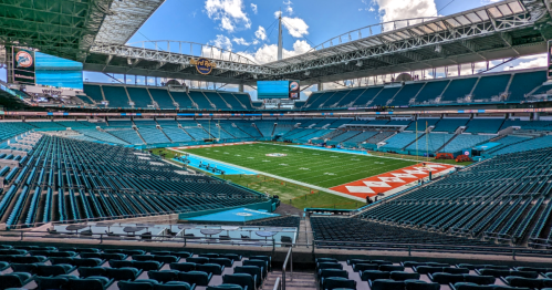 A wide view of a stadium with teal seating and a football field, under a bright blue sky with scattered clouds.