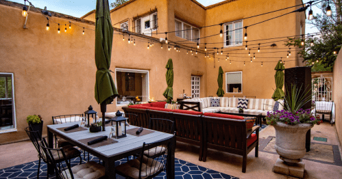 A cozy outdoor patio with string lights, seating, and a dining table surrounded by greenery and warm-colored walls.