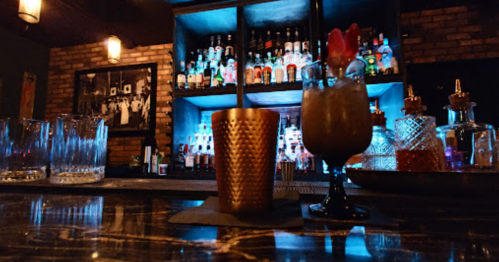 A dimly lit bar scene featuring two cocktails in front of a colorful backlit shelf of liquor bottles.