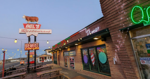 A restaurant exterior with a cow sign, neon lights, and outdoor seating, set against a twilight sky.