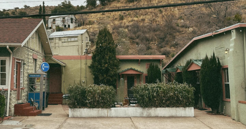 A quaint building complex with green and stone exteriors, surrounded by shrubs and hills in the background.