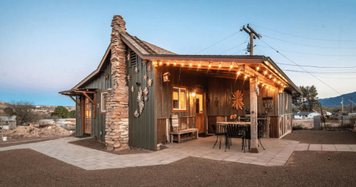 Cozy rustic house with stone accents, string lights, and outdoor seating in a serene setting at dusk.