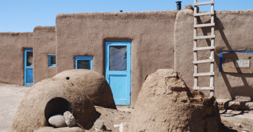 Adobe-style buildings with blue doors and a wooden ladder against a clear blue sky. Earth mounds in the foreground.