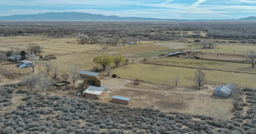 Aerial view of a rural landscape with fields, trees, and several buildings surrounded by mountains in the distance.