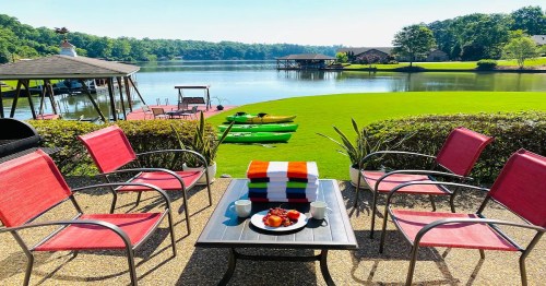 A serene lakeside view with red chairs, a table of fruit, and kayaks on the water, surrounded by lush greenery.