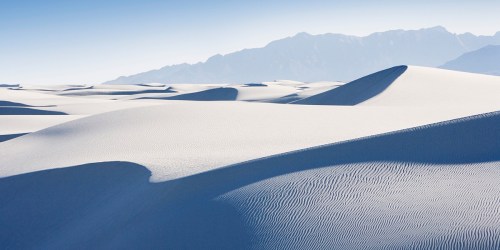 A vast expanse of white sand dunes under a clear blue sky, with distant mountains in the background.