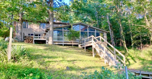 A modern cabin with large windows, surrounded by trees, on a grassy hillside with wooden steps leading up to it.