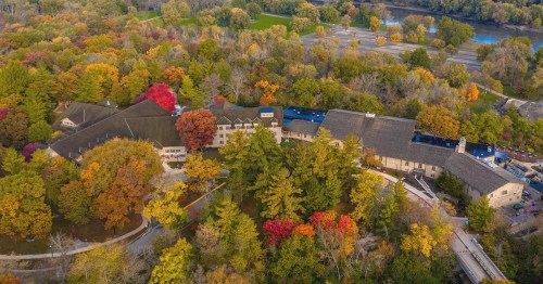 Aerial view of a lodge surrounded by vibrant autumn foliage and a winding path, near a river and green fields.