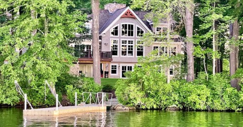 A large, modern house with many windows, surrounded by trees, next to a calm lake and a small wooden dock.