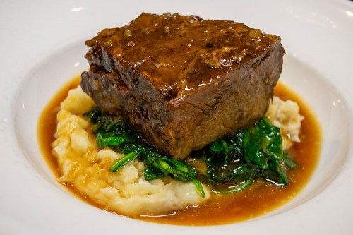 A serving of tender beef on a bed of creamy mashed potatoes, topped with sautéed greens and a rich gravy.