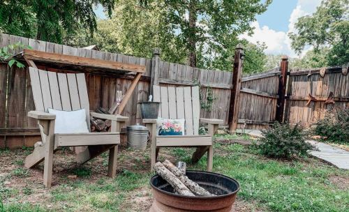 Two wooden chairs with cushions sit beside a fire pit in a backyard, surrounded by a wooden fence and greenery.
