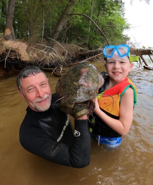 A man and a child in water, holding a large catfish, both smiling and wearing snorkeling gear.
