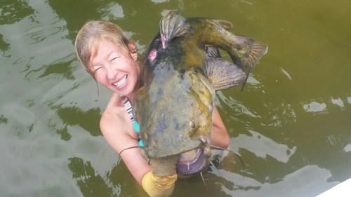 A smiling woman in water holds a large catfish, showcasing her catch with joy.