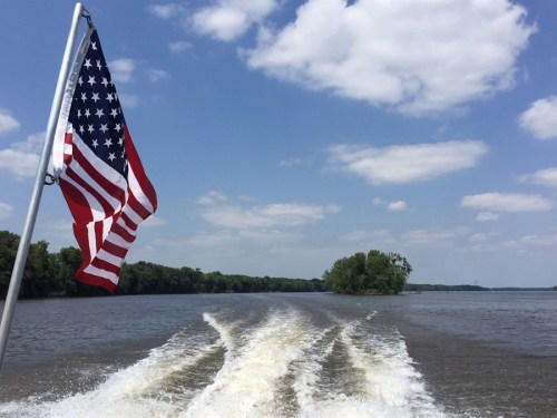 A boat's wake trails behind as an American flag waves in the breeze against a backdrop of blue sky and green trees.