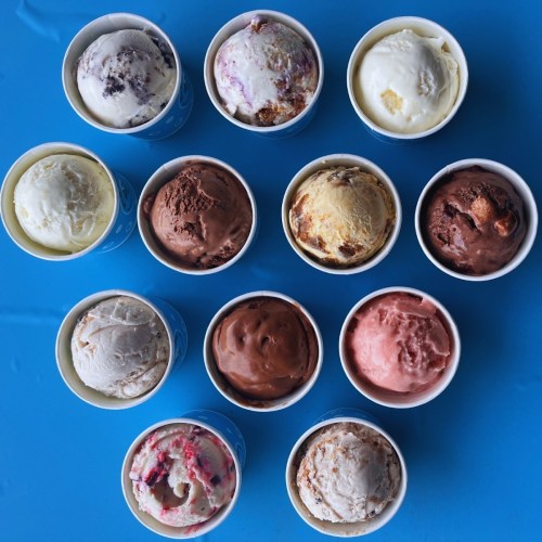 Twelve cups of various ice cream flavors arranged in a grid on a blue surface.