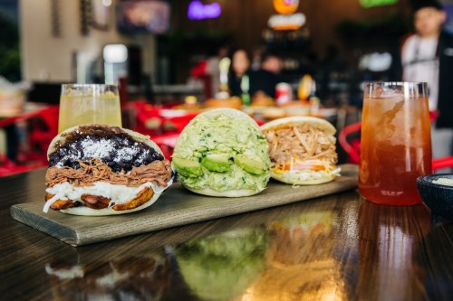 Three flavorful bao buns filled with pulled pork, avocado, and shredded chicken, served on a wooden platter with drinks.