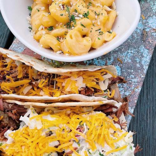 A wooden platter with three tacos filled with shredded meat and cheese, alongside a bowl of creamy macaroni and cheese.