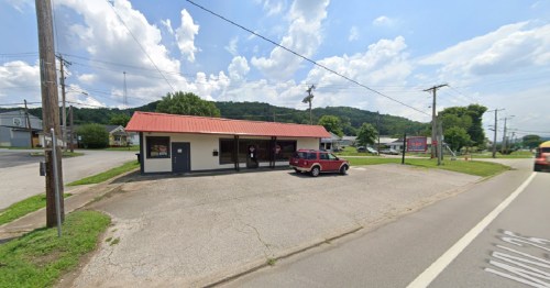 A small commercial building with a red roof, surrounded by greenery and power lines, along a rural road.