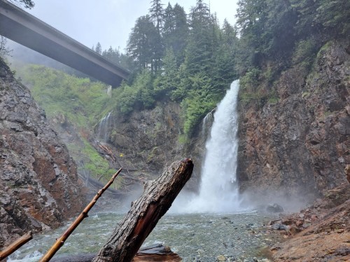 A waterfall cascades down rocky cliffs, surrounded by lush greenery, with a bridge overhead and mist rising from the water.