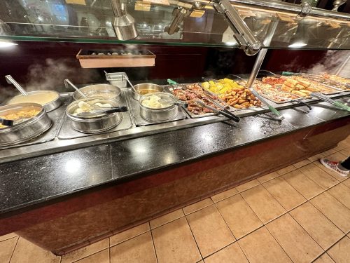 A buffet with various dishes in metal trays, including meats, vegetables, and sauces, on a black countertop.