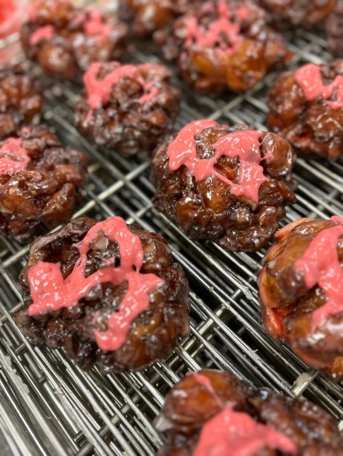 A close-up of glazed pastries with a shiny chocolate coating and pink icing drizzled on top, arranged on a cooling rack.