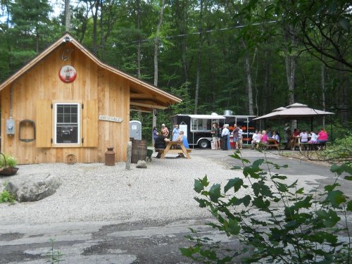 A wooden cabin with a food truck nearby, surrounded by trees and people dining under umbrellas.
