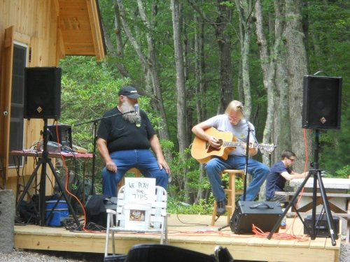 Two musicians perform on an outdoor stage surrounded by trees, with one playing guitar and the other seated nearby.