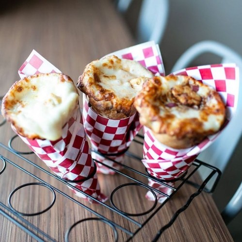 Three cone-shaped servings of cheesy baked dishes in red and white checkered paper, displayed on a wire rack.