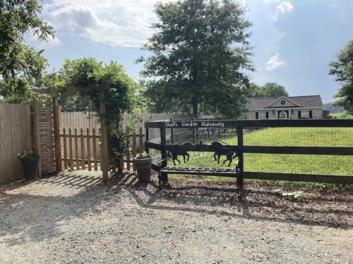 A wooden gate with a horse silhouette leads to a garden, surrounded by trees and a house in the background.