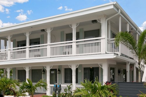 A two-story white building with balconies, surrounded by greenery and blue skies.