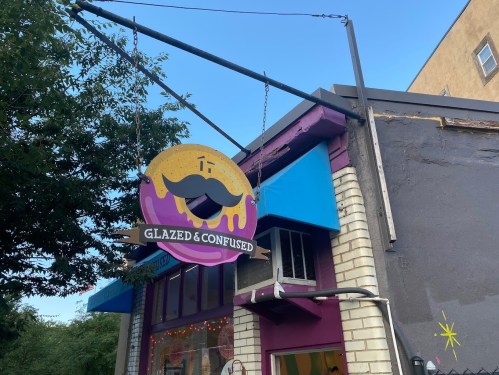 Sign for "Glazed & Confused" featuring a donut with a mustache, hanging from a building with colorful awnings.