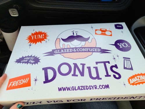 A colorful donut box labeled "Glazed and Confused Donuts" with playful graphics and text, resting in a car.
