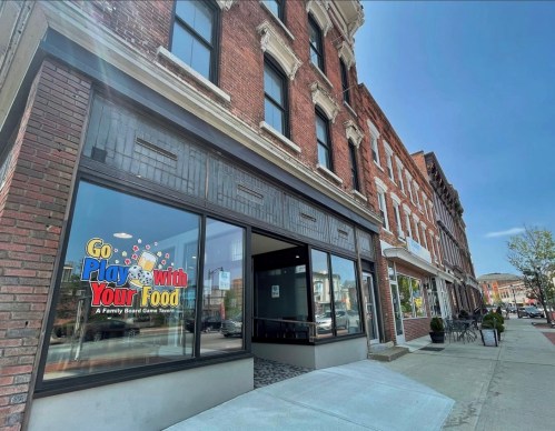 Exterior of a brick building featuring a colorful sign for "Go Play with Your Food," a family board game store.