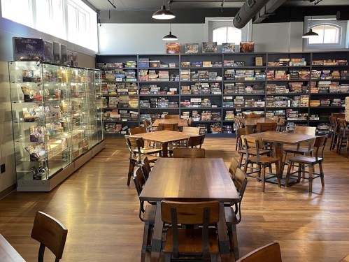A cozy game café with wooden tables, chairs, and shelves filled with board games and collectibles.