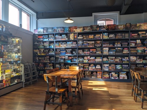 A cozy game store interior with a wooden table, chairs, and shelves filled with various board games.