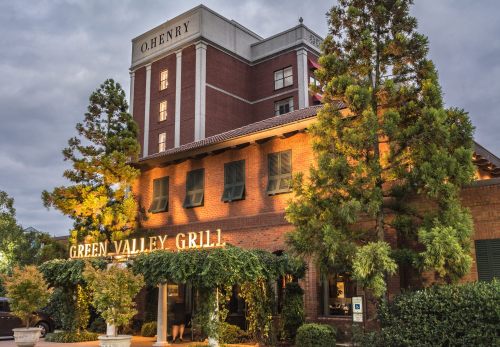 Exterior view of Green Valley Grill with lush greenery, illuminated sign, and O. Henry hotel in the background.