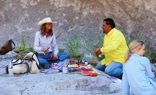 Two people sit on the ground sharing a meal, while a third person sits nearby, all surrounded by nature.