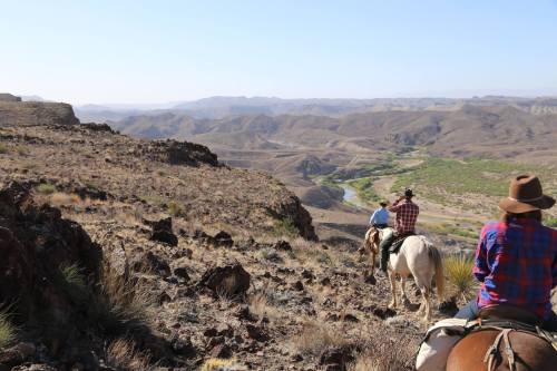 Two horseback riders navigate a rocky terrain with a vast, arid landscape and winding river in the background.